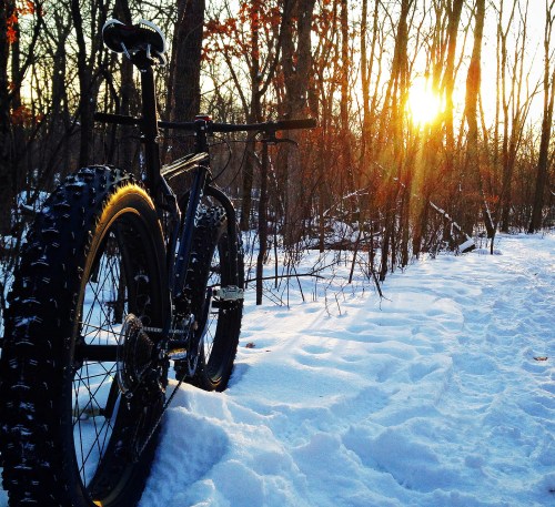 A fat bike rests on a snowy trail as the sun sets behind trees, casting a warm glow in the winter landscape.