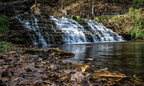 A serene waterfall cascades over rocks into a calm pool, surrounded by autumn leaves and lush greenery.