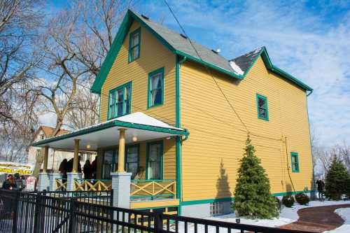 A yellow two-story house with green trim, surrounded by snow and trees, under a blue sky with scattered clouds.