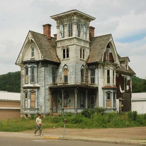 A dilapidated Victorian house with peeling blue paint, surrounded by overgrown grass and a person walking nearby.