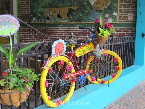 A colorful bicycle adorned with flowers, parked against a brick wall with greenery and decorative elements.