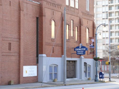 Exterior of Ebenezer Baptist Church, featuring brick walls, arched windows, and a blue sign. Urban setting with nearby buildings.