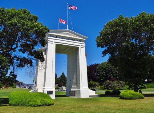 White archway with flags, surrounded by green trees and grass, inscribed "Children of a Common Mother." Clear blue sky.