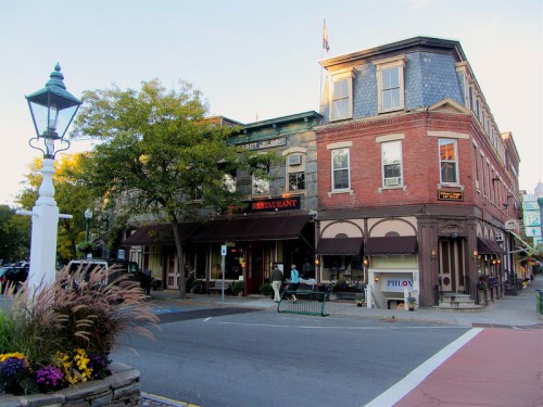 Historic brick buildings with a restaurant sign, surrounded by trees and flowers, in a quaint town setting.