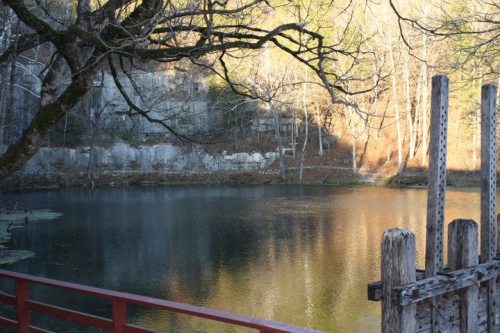 A serene pond surrounded by trees and rocky cliffs, reflecting warm sunlight in a tranquil setting.