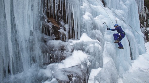 A climber ascends a frozen waterfall using ice axes and crampons, surrounded by icy rock formations.