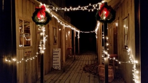 A festive pathway lined with lights and wreaths, leading through a rustic wooden building at night.