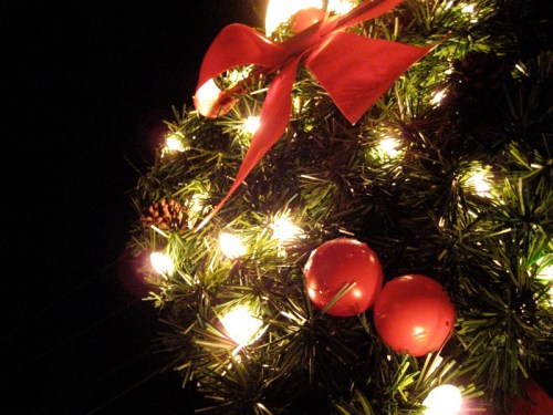 A close-up of a Christmas wreath with red ornaments, a bow, and glowing lights against a dark background.