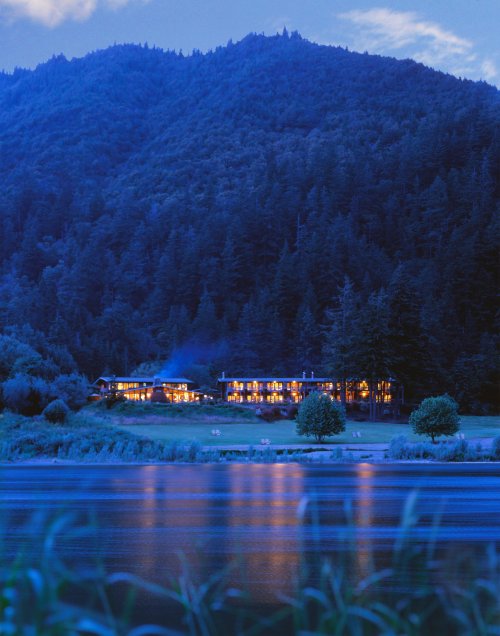 A serene lakeside view at dusk, featuring a cozy lodge nestled among mountains and trees, reflecting in the water.