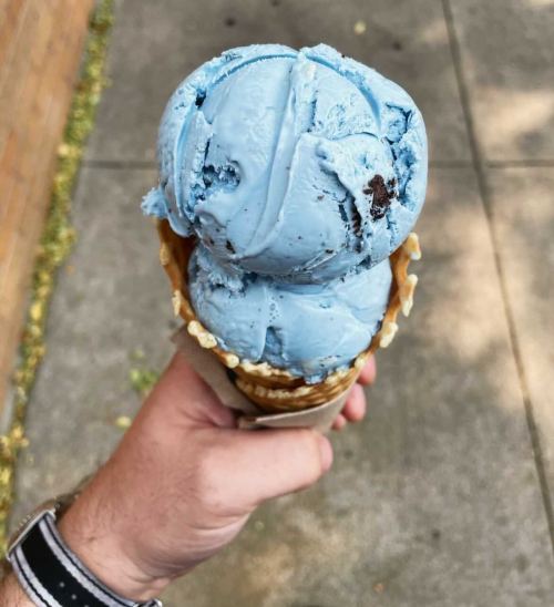 A hand holding a cone with two scoops of blue ice cream, with a sidewalk and brick wall in the background.