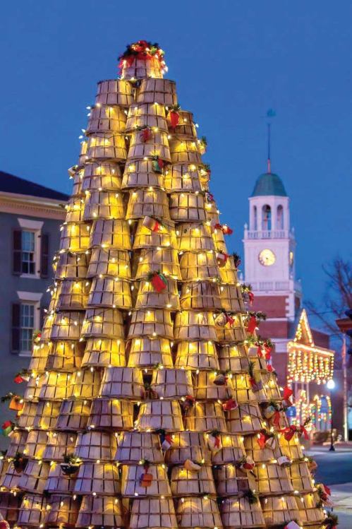 A festive tree made of stacked baskets, adorned with lights and decorations, set against a twilight sky and clock tower.