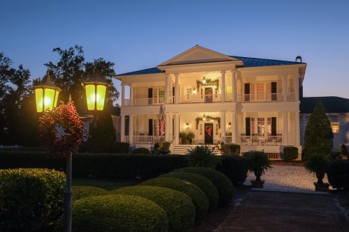 A grand two-story house with white columns, decorated for the holidays, illuminated at dusk with landscaped gardens.