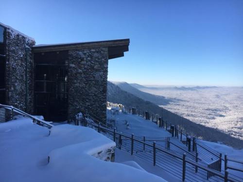 A snowy landscape with a modern stone building overlooking a vast valley and mountains under a clear blue sky.
