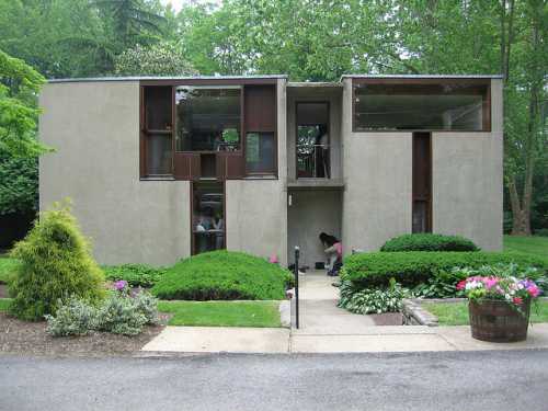 Modern concrete house surrounded by lush greenery and colorful flowers, featuring large windows and a unique design.