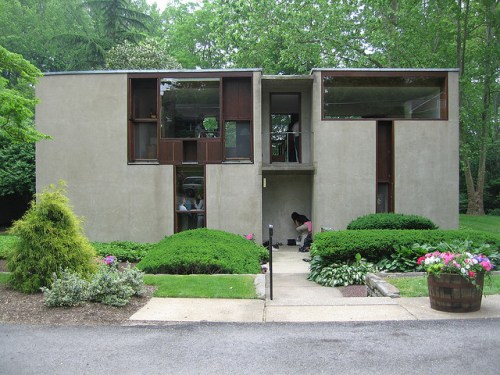 Modern concrete house surrounded by lush greenery and colorful flowers, featuring large windows and a unique design.