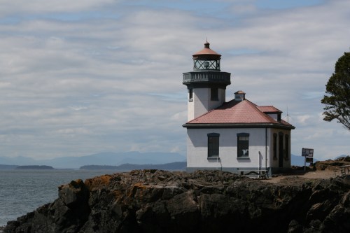 A white lighthouse with a red roof stands on rocky shore, overlooking the ocean under a cloudy sky.