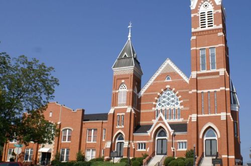 A large red brick church with tall spires and a stained glass window, set against a clear blue sky.