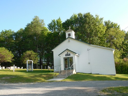 A small white church with a steeple, surrounded by trees and a grassy area, on a clear sunny day.
