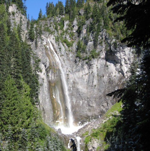 A tall waterfall cascading down a rocky cliff, surrounded by lush green trees and a clear blue sky.