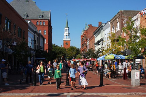 A bustling street scene with people walking, shops lining the street, and a church steeple in the background.