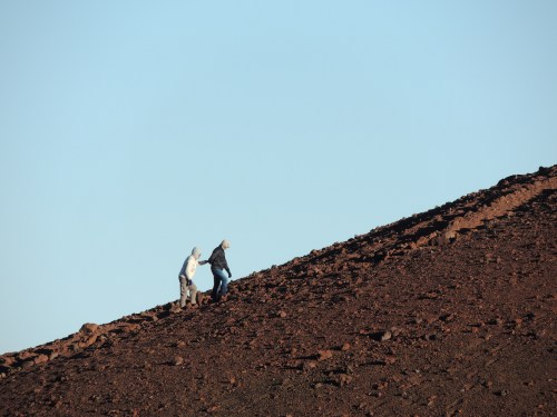 Two people climb a rocky, sloped terrain against a clear blue sky.