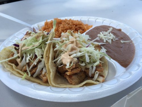 Two tacos filled with grilled fish, topped with cabbage and sauce, served with rice and refried beans on a plate.