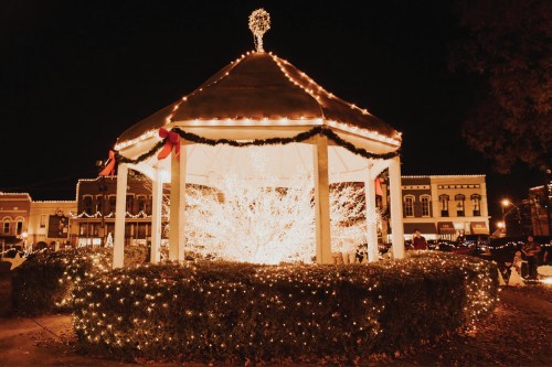 A beautifully lit gazebo surrounded by festive lights, set against a night sky in a charming town square.