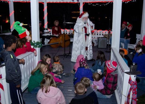 A festive gathering with Santa in a decorated gazebo, surrounded by children and musicians, celebrating the holiday season.