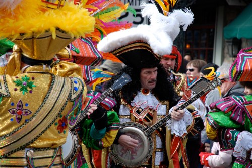 Colorful performers in elaborate costumes play instruments at a festive parade, surrounded by an enthusiastic crowd.