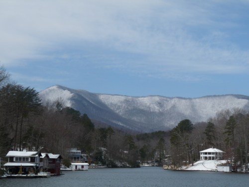 Snow-covered mountains rise above a serene lake, with cozy houses lining the shore under a clear blue sky.