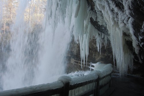 Icicles hang from a rocky overhang near a frozen waterfall, with a wooden railing in the foreground.