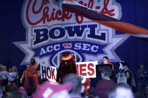 A mascot and fans at a sports event, holding signs in front of a backdrop for the Chick-fil-A Bowl.