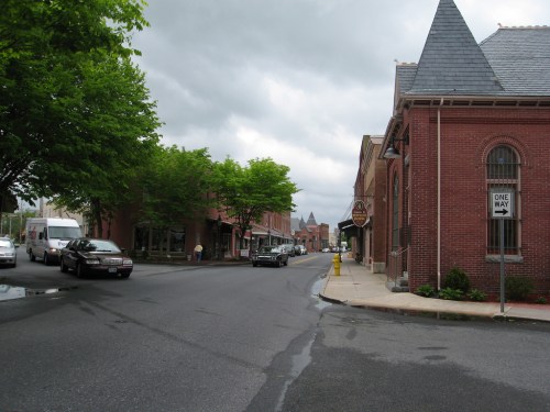 A quiet street scene with brick buildings, trees, and cloudy skies, featuring parked cars and a one-way sign.