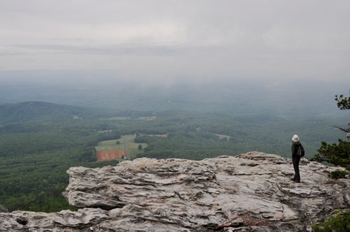 A person stands on a rocky ledge, gazing out over a misty green valley and distant hills.