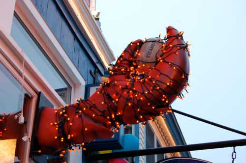 A large, red lobster claw adorned with colorful string lights, hanging outside a building.