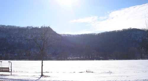 A snowy landscape with a river, bare trees, and hills under a bright blue sky.
