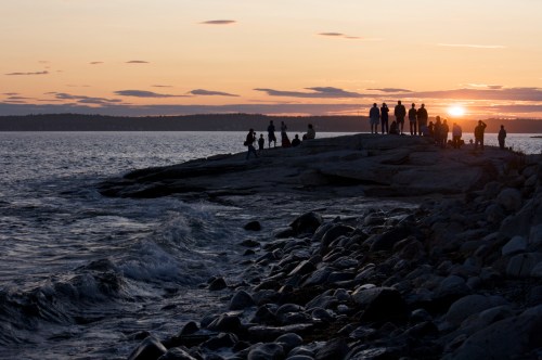 Silhouettes of people on a rocky shore at sunset, with waves gently crashing against the rocks.