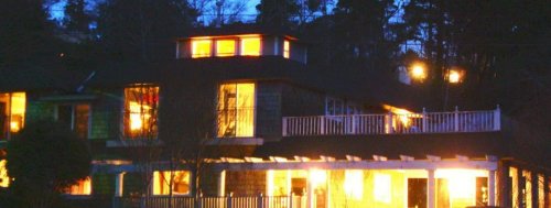 A cozy house at night, illuminated from within, surrounded by trees and a porch with a railing.