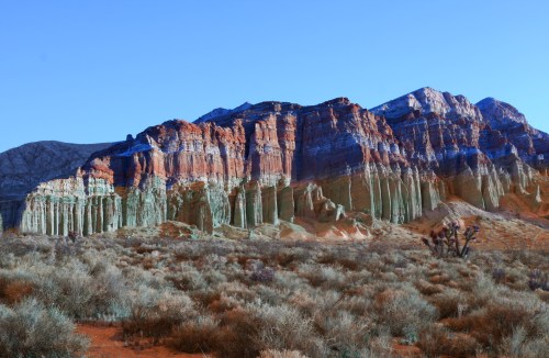 Colorful rock formations rise against a clear blue sky, surrounded by desert vegetation and rugged terrain.
