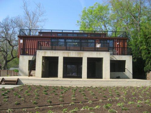 A modern two-story building with a concrete base and a metal upper structure, surrounded by greenery and a clear blue sky.
