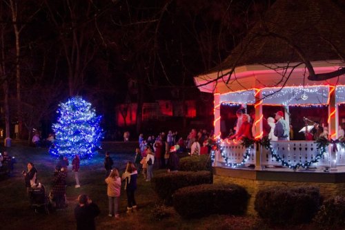 A festive scene at night with a decorated gazebo, a blue Christmas tree, and a crowd enjoying holiday festivities.