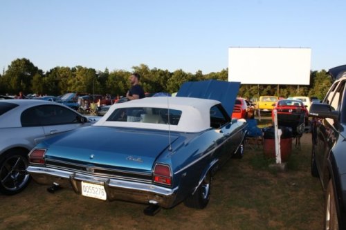 A classic blue convertible car parked at a gathering, with a man standing nearby and a blank billboard in the background.