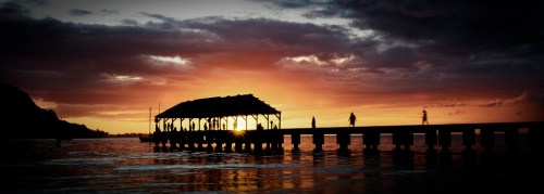 Silhouette of a pier at sunset, with people walking and vibrant colors in the sky reflecting on the water.