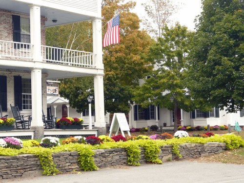 A charming building with a porch, American flag, and colorful flower beds in a serene outdoor setting.