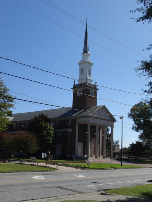 A tall church with a steeple, surrounded by trees and power lines, under a clear blue sky.