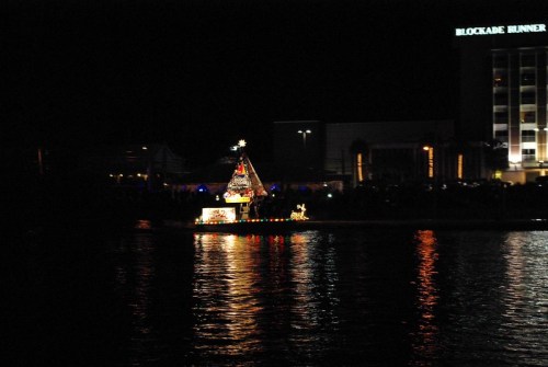 A brightly lit Christmas tree boat floats on dark water, with a hotel and crowd visible in the background.