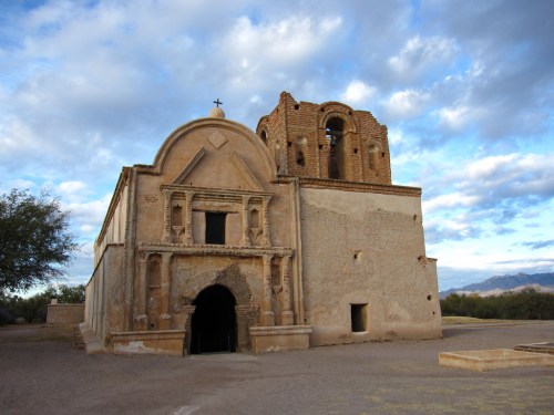 Historic adobe church with a bell tower, set against a cloudy sky and desert landscape.