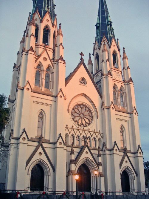 A tall, ornate church with two spires, intricate architecture, and a large rose window, illuminated by soft evening light.