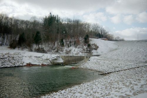 A snowy landscape featuring a dam, a calm water body, and trees in the background under a cloudy sky.