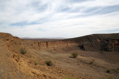 A barren landscape with rocky terrain, sparse vegetation, and distant mountains under a cloudy sky.